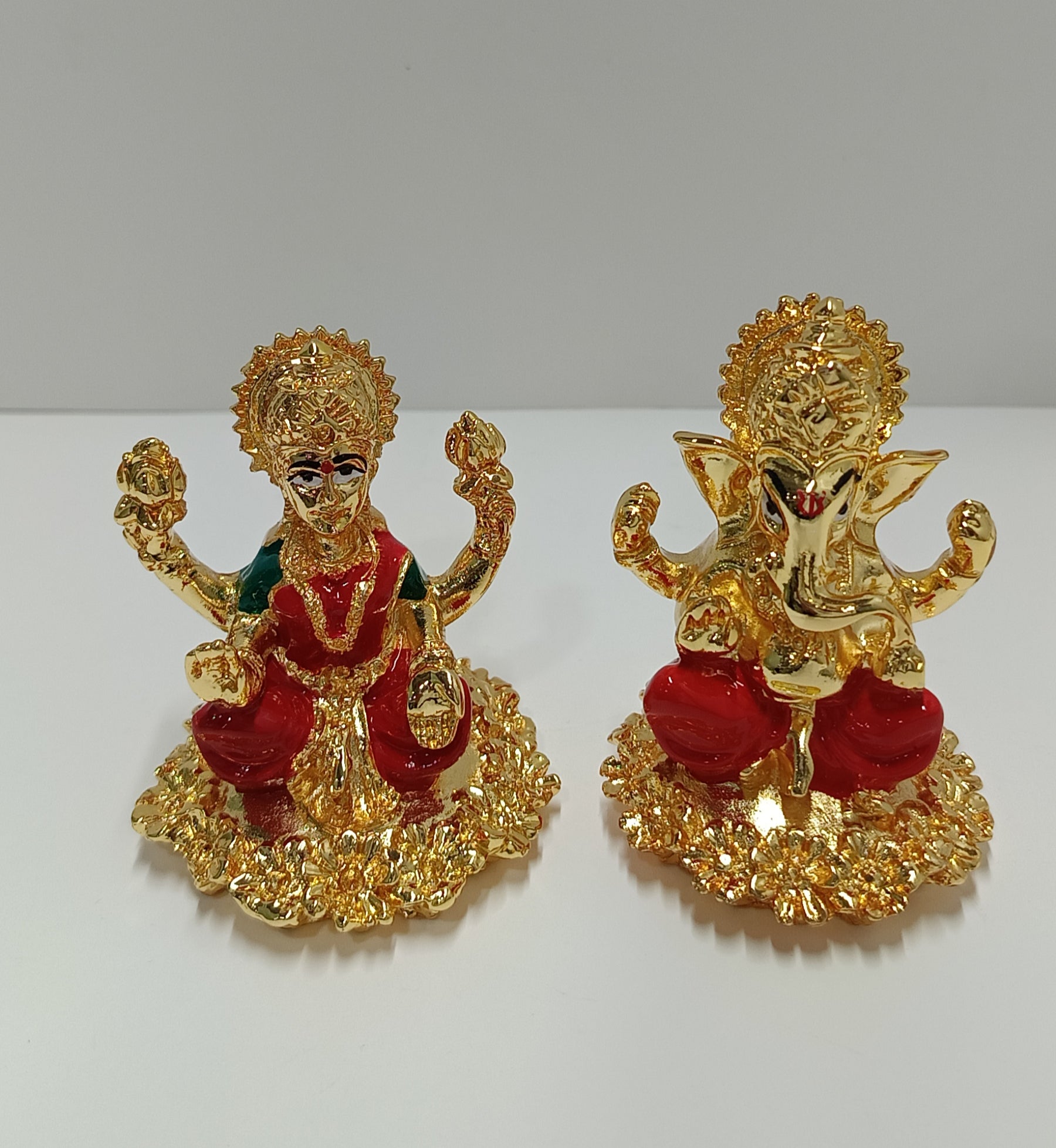 A pair of polyresin gold-plated statues depicting Goddess Lakshmi and Lord Ganesha, seated on elaborate golden bases with red and green accents on their attire.