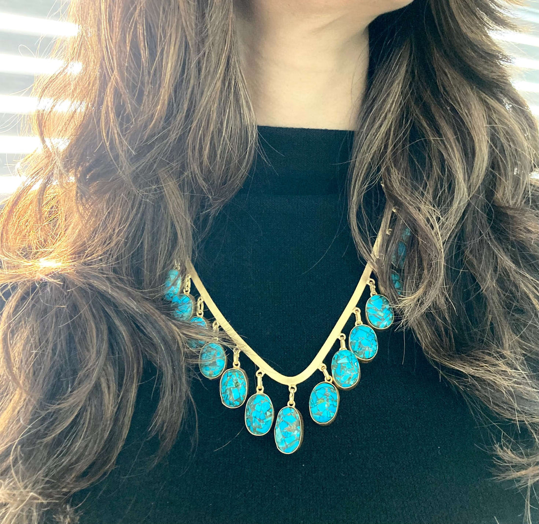 A woman wearing a black top and a V-shaped necklace with blue beads and gold accents.