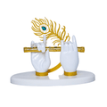 A white statue depicting Lord Krishna's hands, one holding a gold flute and the other in a gesture, adorned with a sparkling gold and blue peacock feather, all resting on a simple white base.