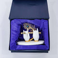 A decorative figurine depicting Lord Krishna's white hands holding a golden flute, adorned with a golden peacock feather, displayed inside a dark box lined with royal blue fabric