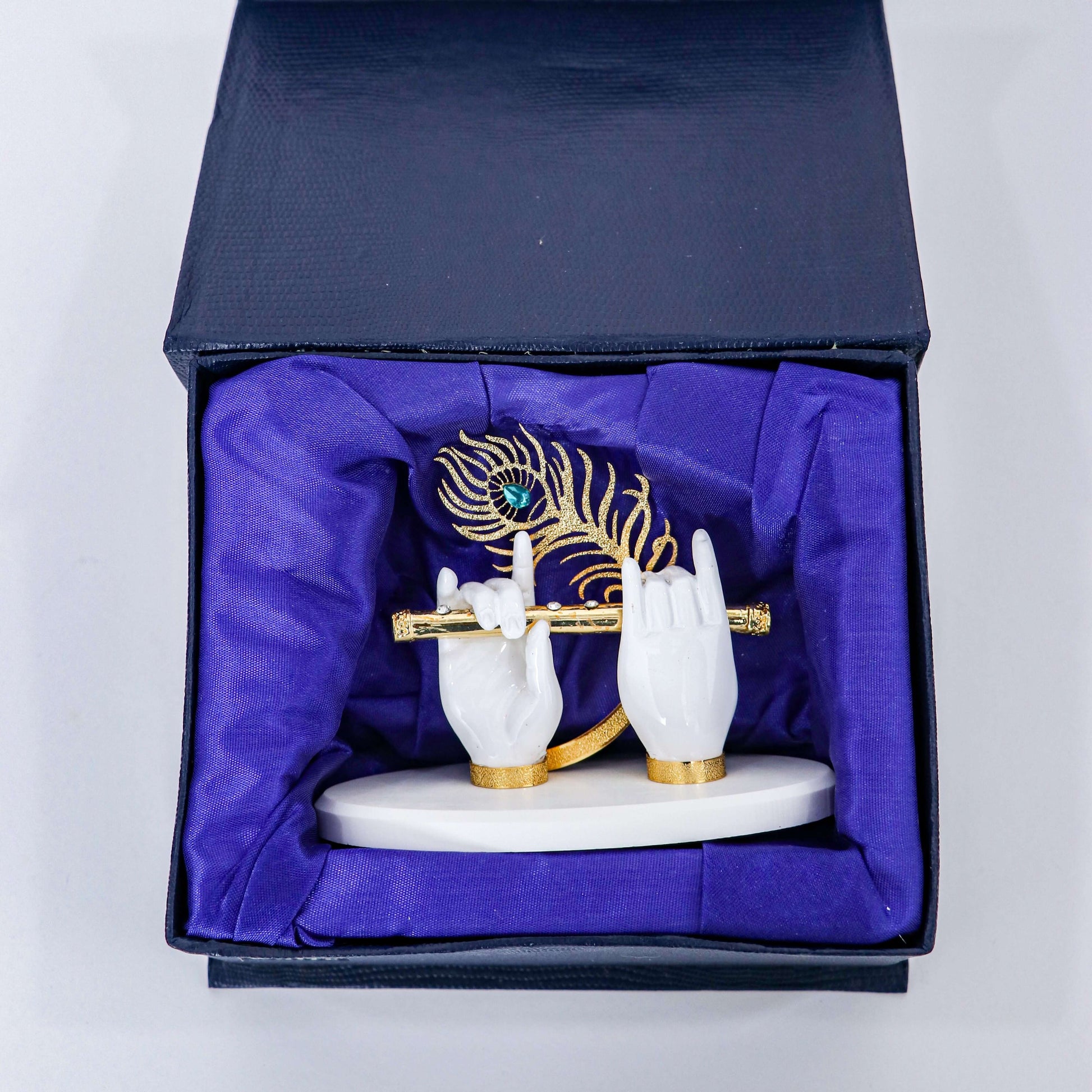 A decorative figurine depicting Lord Krishna's white hands holding a golden flute, adorned with a golden peacock feather, displayed inside a dark box lined with royal blue fabric
