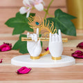 A decorative showpiece featuring two white sculpted hands, representing Lord Krishna's hands, gracefully holding a golden flute. A golden peacock feather with a blue jewel accent is positioned above the hands. The sculpture rests on a white, round base.