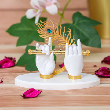 A decorative showpiece featuring two white sculpted hands, representing Lord Krishna's hands, gracefully holding a golden flute. A golden peacock feather with a blue jewel accent is positioned above the hands. The sculpture rests on a white, round base.