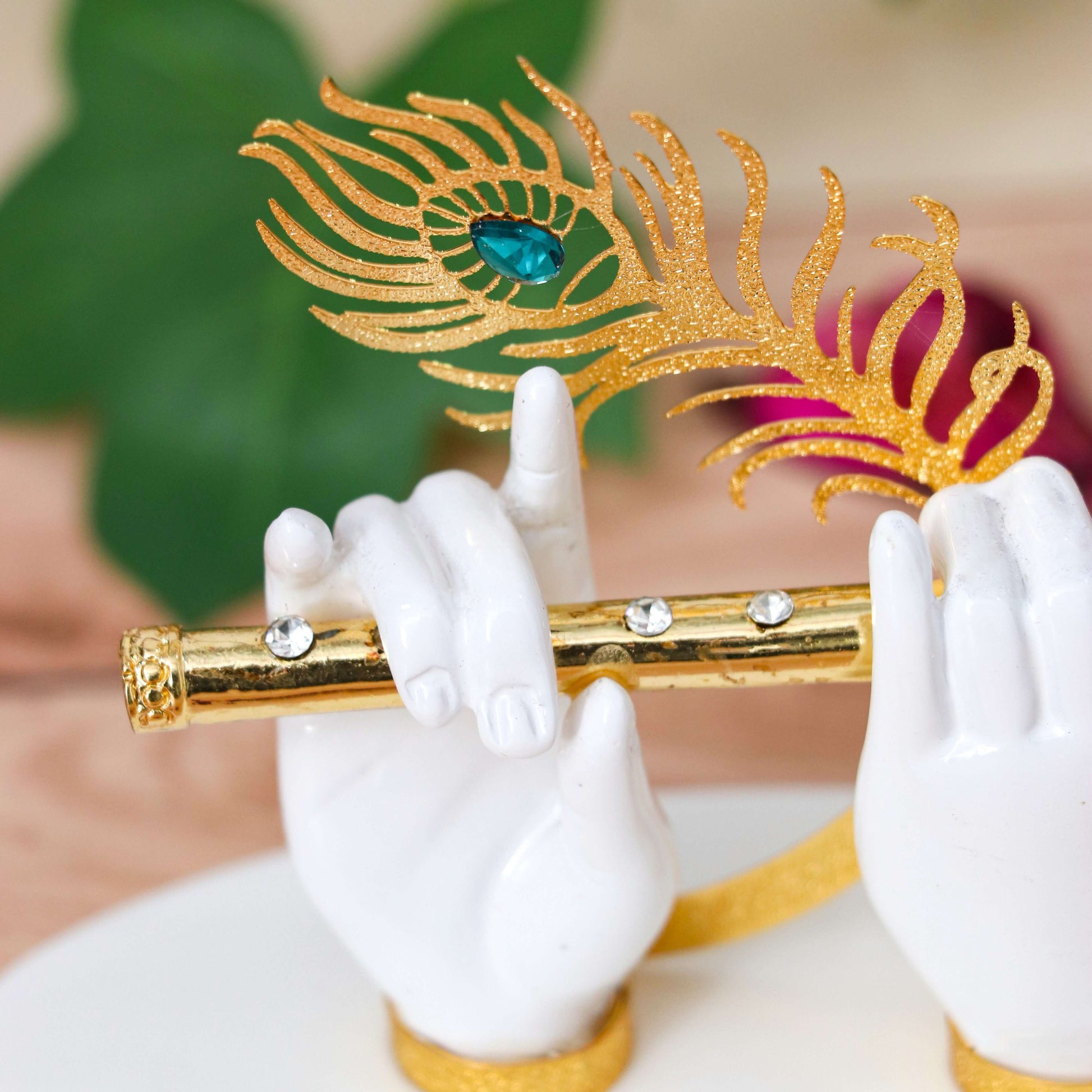 A decorative idol featuring white hands, likely representing Lord Krishna, holding a golden flute adorned with clear gems, and a golden peacock feather with a prominent blue gemstone. The background is softly blurred green foliage.