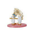 A decorative idol featuring Lord Krishna's hands in white holding a golden flute, with a golden peacock feather adorned with a blue eye-spot behind them, all mounted on a polished pink base.