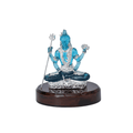 A blue crystal or glass idol of Lord Shiva in a meditative pose (Padmasana) on a silver-colored pedestal, holding a trishul (trident) in one hand and another object in a raised hand, all mounted on a dark brown, polished wooden base. The idol features intricate details like Shiva's third eye, snake around the neck, and traditional attire.