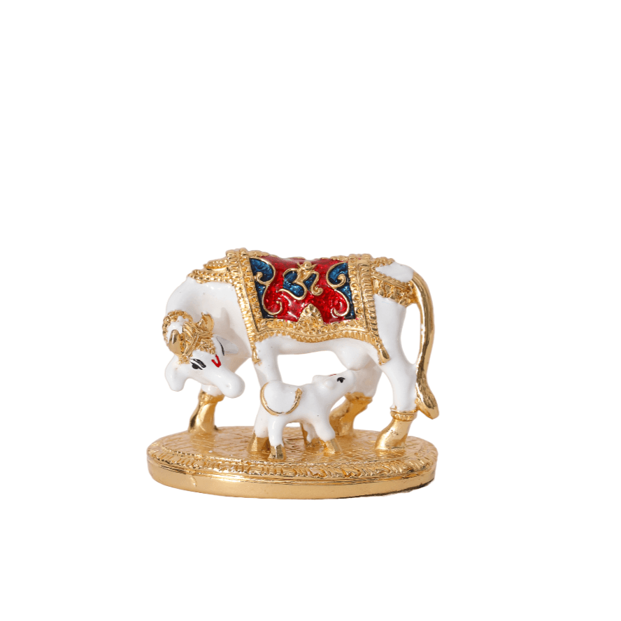 A golden and white statue of a Kamadhenu cow with her calf, adorned with intricate red and blue details on the cow's back, standing on a circular base