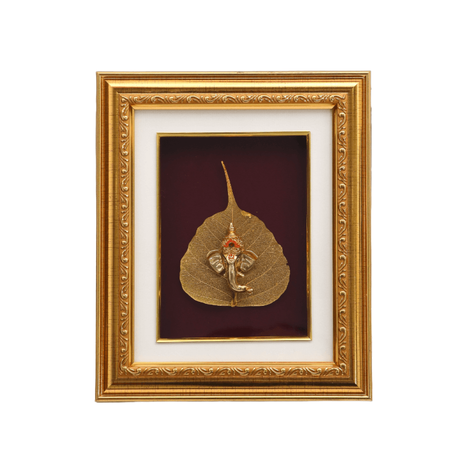 Framed artwork depicting a small, metallic Lord Ganesha idol resting on a golden or gilded Peepal leaf, set against a deep red background within an ornate gold picture frame.
