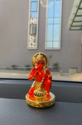 A vibrant orange-red and gold Lord Hanuman car dashboard idol is shown kneeling on a golden, ornate base. The deity is holding a golden mace (gada) in one hand and raising the other in a gesture of blessing.