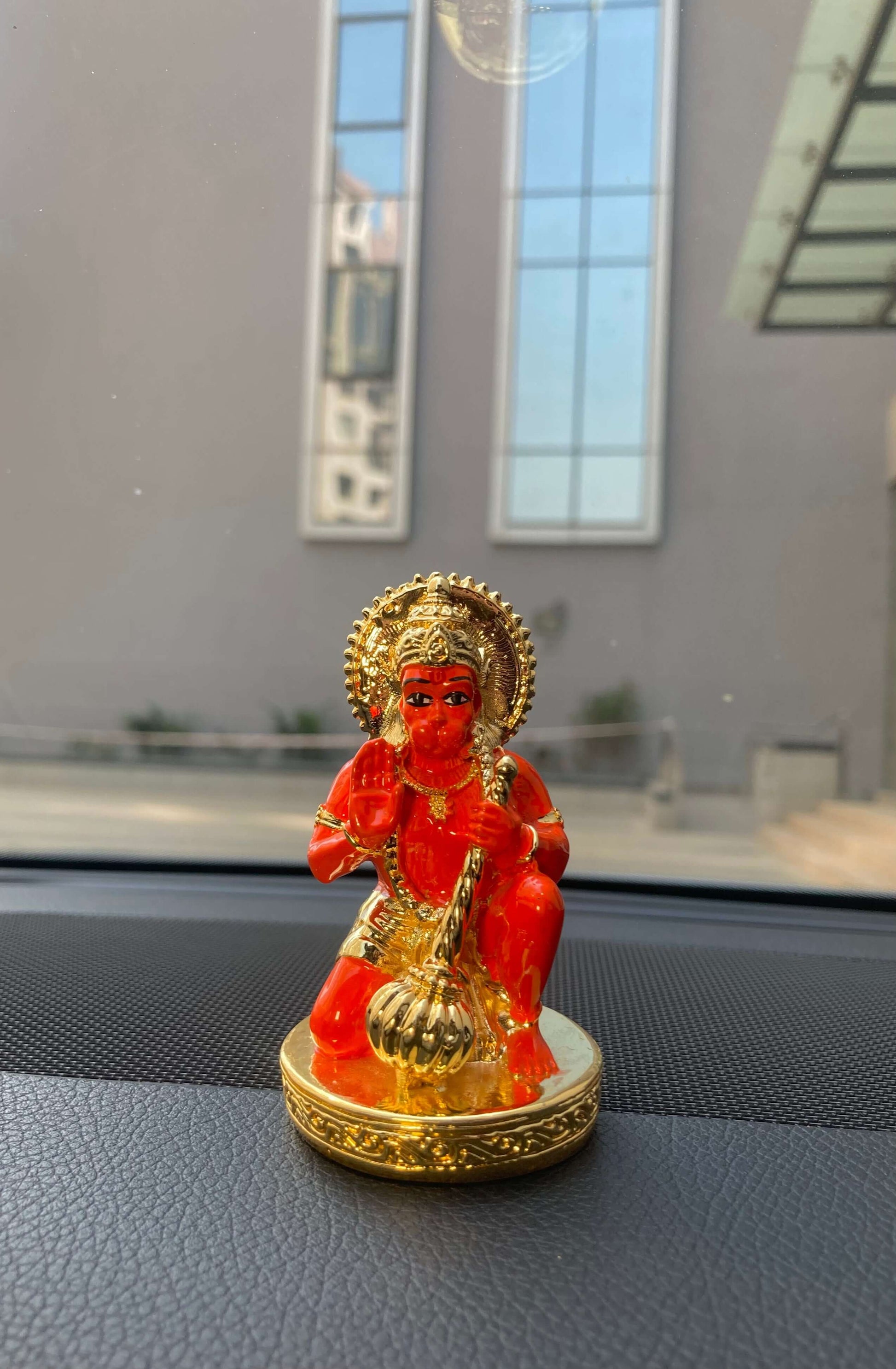 A vibrant orange-red and gold Lord Hanuman car dashboard idol is shown kneeling on a golden, ornate base. The deity is holding a golden mace (gada) in one hand and raising the other in a gesture of blessing.