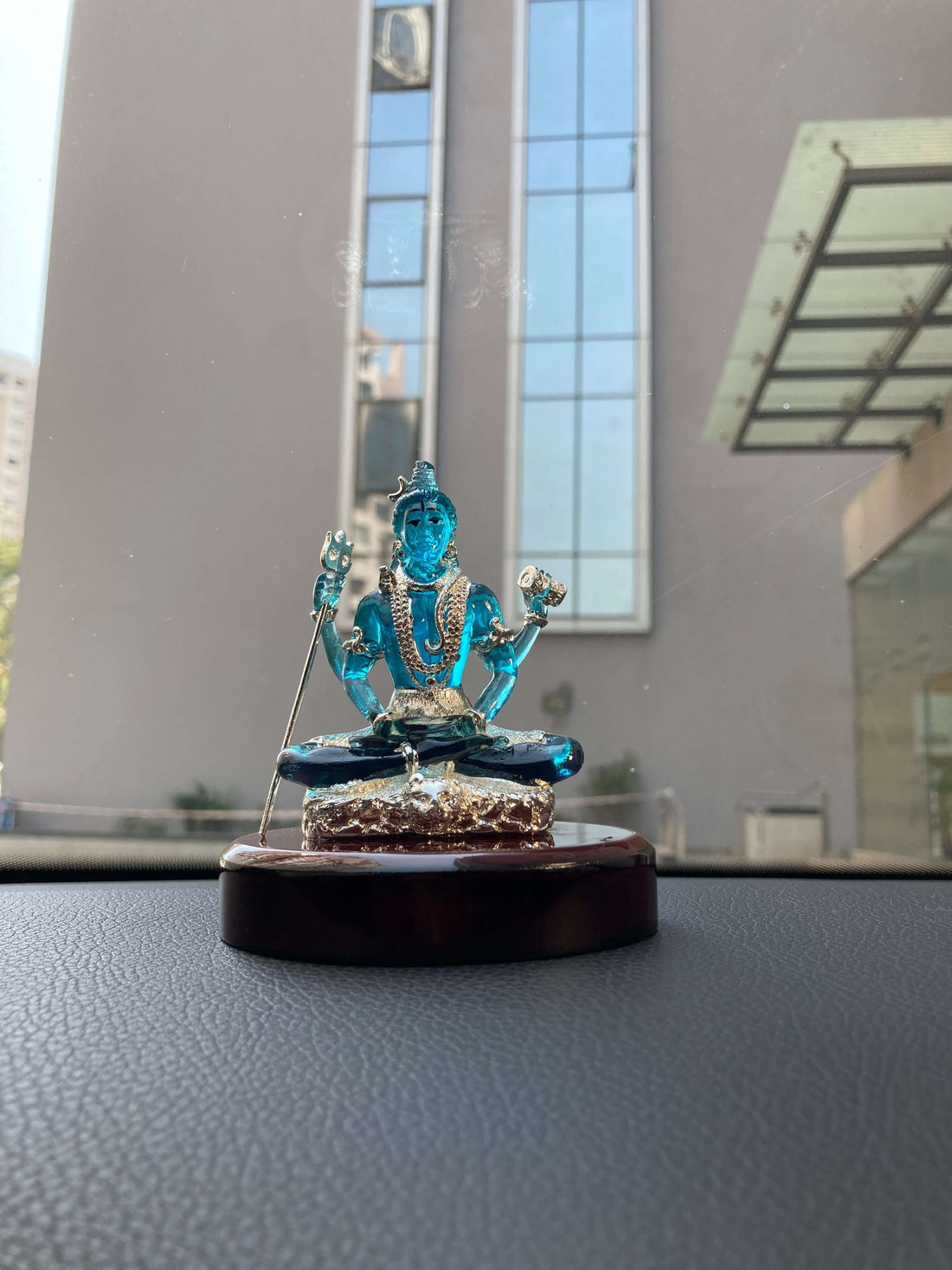 A close-up of a vibrant blue, translucent Lord Shiva idol, made crystal, adorned with intricate silver-colored details, seated in a meditative pose on a polished dark wooden base. The background is softly blurred, showing a hint of greenery and rustic elements like pinecones.