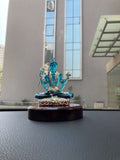 A blue, translucent crystal or glass idol of Lord Shiva, seated in meditation pose and holding a trident and other attributes, displayed on a dark base, likely on a car dashboard, with a modern building visible in the blurry background.