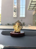 A white and gold Ganesha idol with a detailed Kalpavriksha tree halo, mounted on a glossy red-brown wooden base, is shown on a car's dashboard.