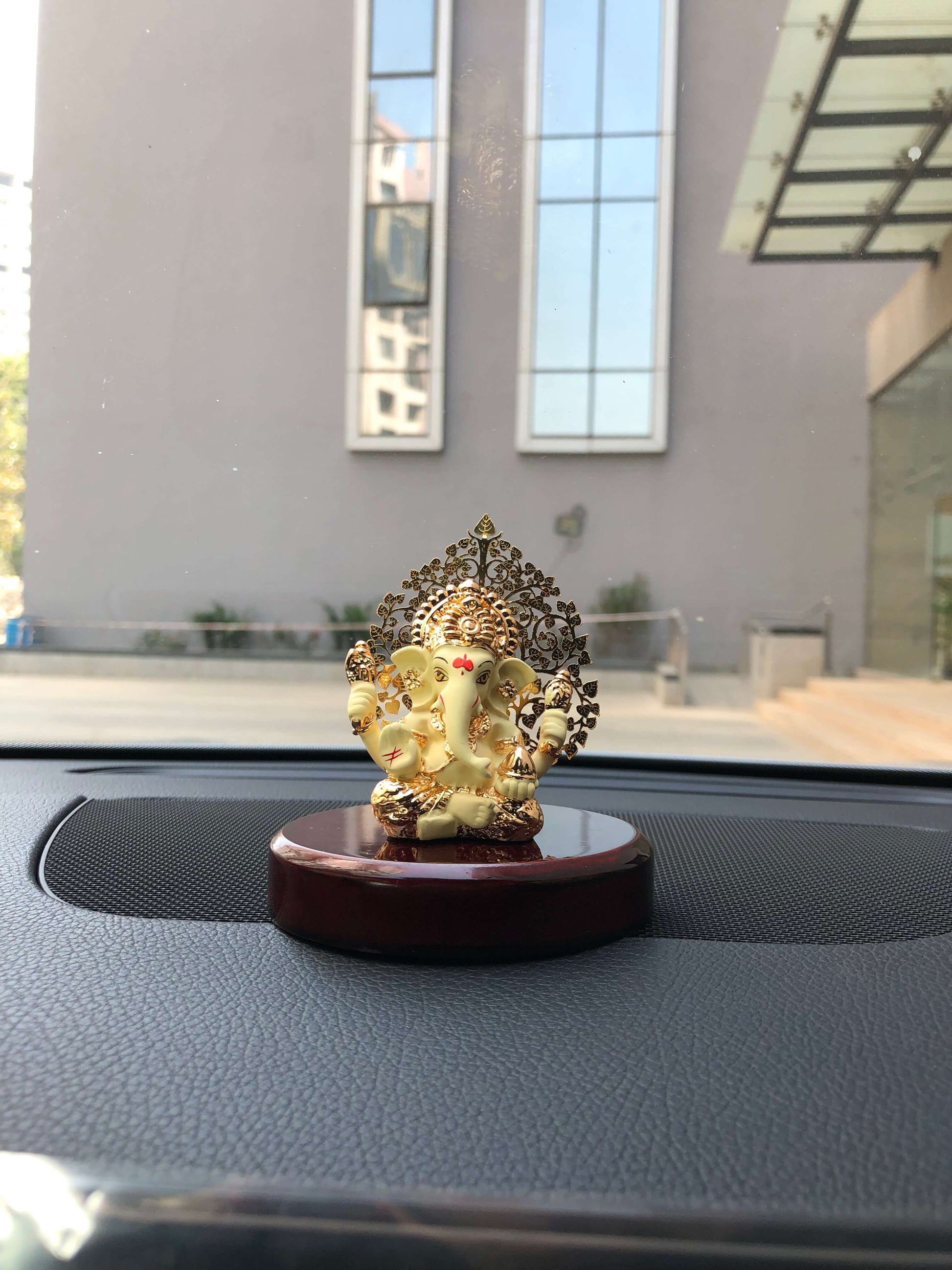 A white and gold Ganesha idol with a detailed Kalpavriksha tree halo, mounted on a glossy red-brown wooden base, is shown on a car's dashboard.