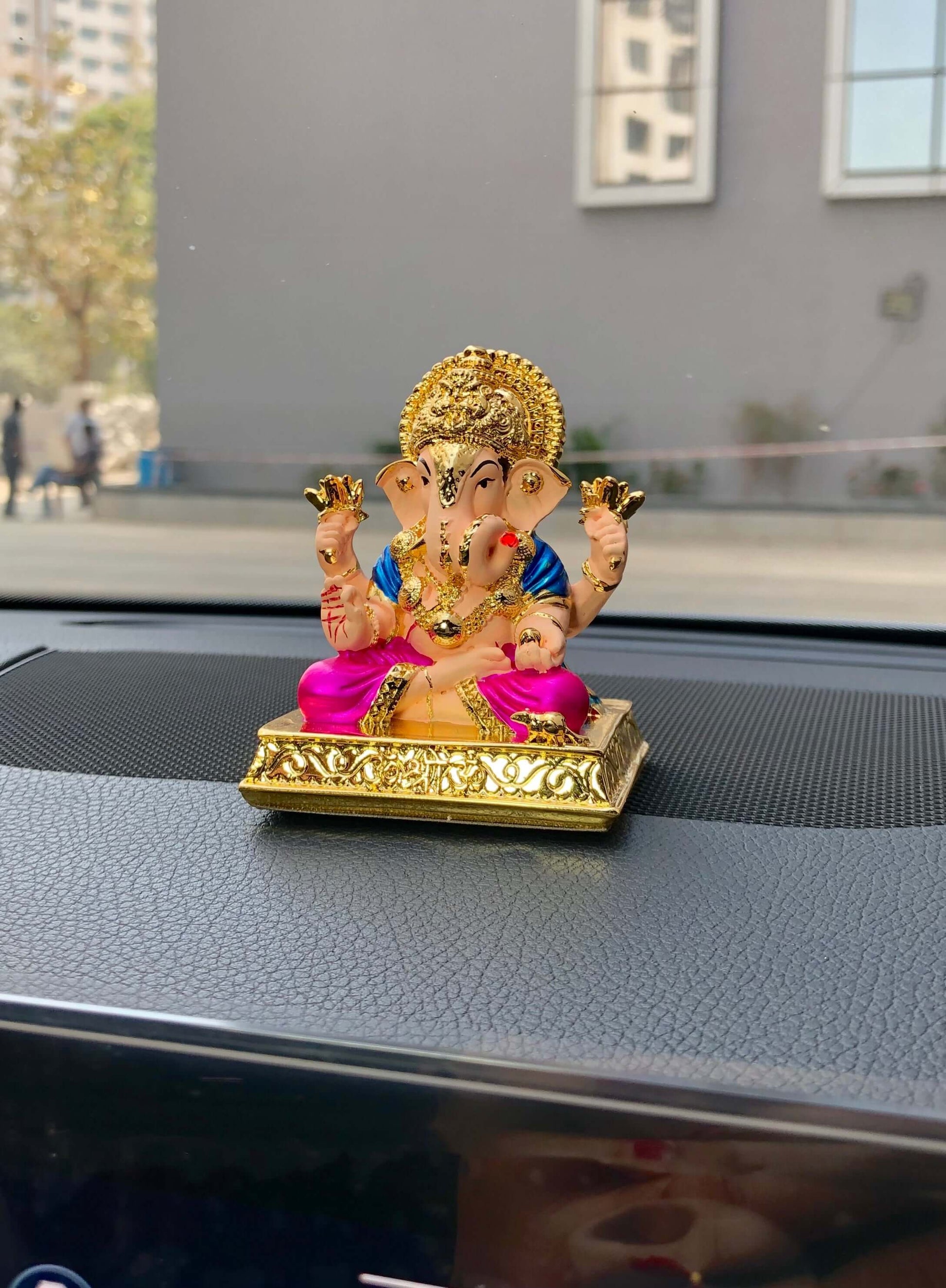 A colorful Dagdushet Ganesha idol with a gold crown and halo, a blue top, and a shiny magenta dhoti, is shown seated on an ornate golden base on a car dashboard.