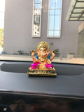 colorful Dagdushet Ganesha idol with a gold crown, a blue top, and a magenta lower garment, seated on an ornate golden base, positioned on a car dashboard.