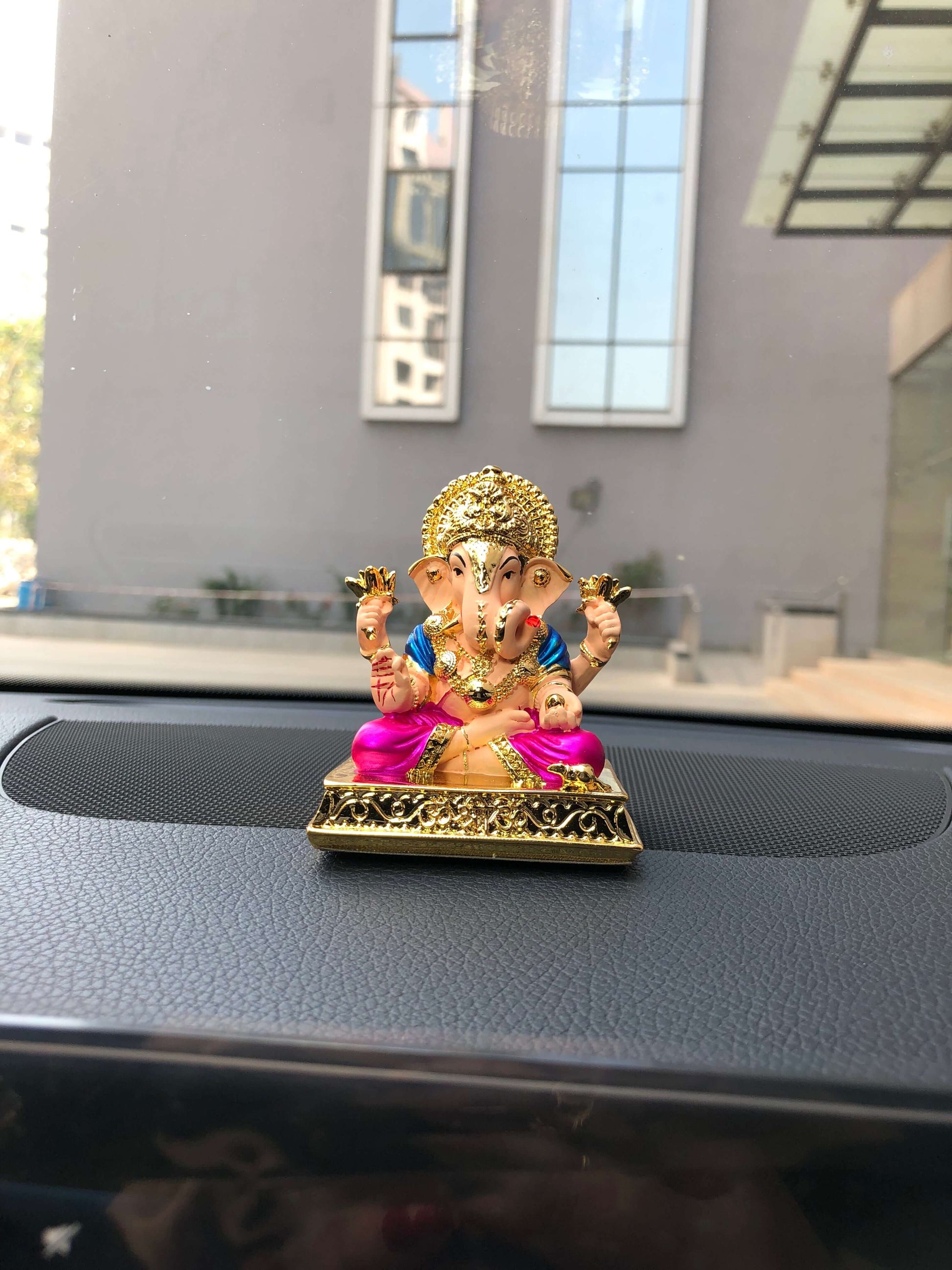 colorful Dagdushet Ganesha idol with a gold crown, a blue top, and a magenta lower garment, seated on an ornate golden base, positioned on a car dashboard.