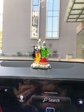 A small, colorful idol of Radha Krishna is placed on the black dashboard of a car, with a blurry background showing a building through the windshield and the car's infotainment screen partially visible below with a 'Search' button.