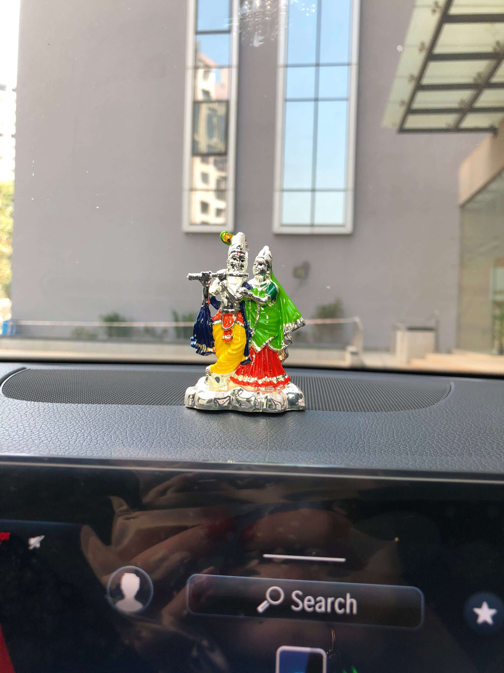 A small, colorful idol of Radha Krishna is placed on the black dashboard of a car, with a blurry background showing a building through the windshield and the car's infotainment screen partially visible below with a 'Search' button.