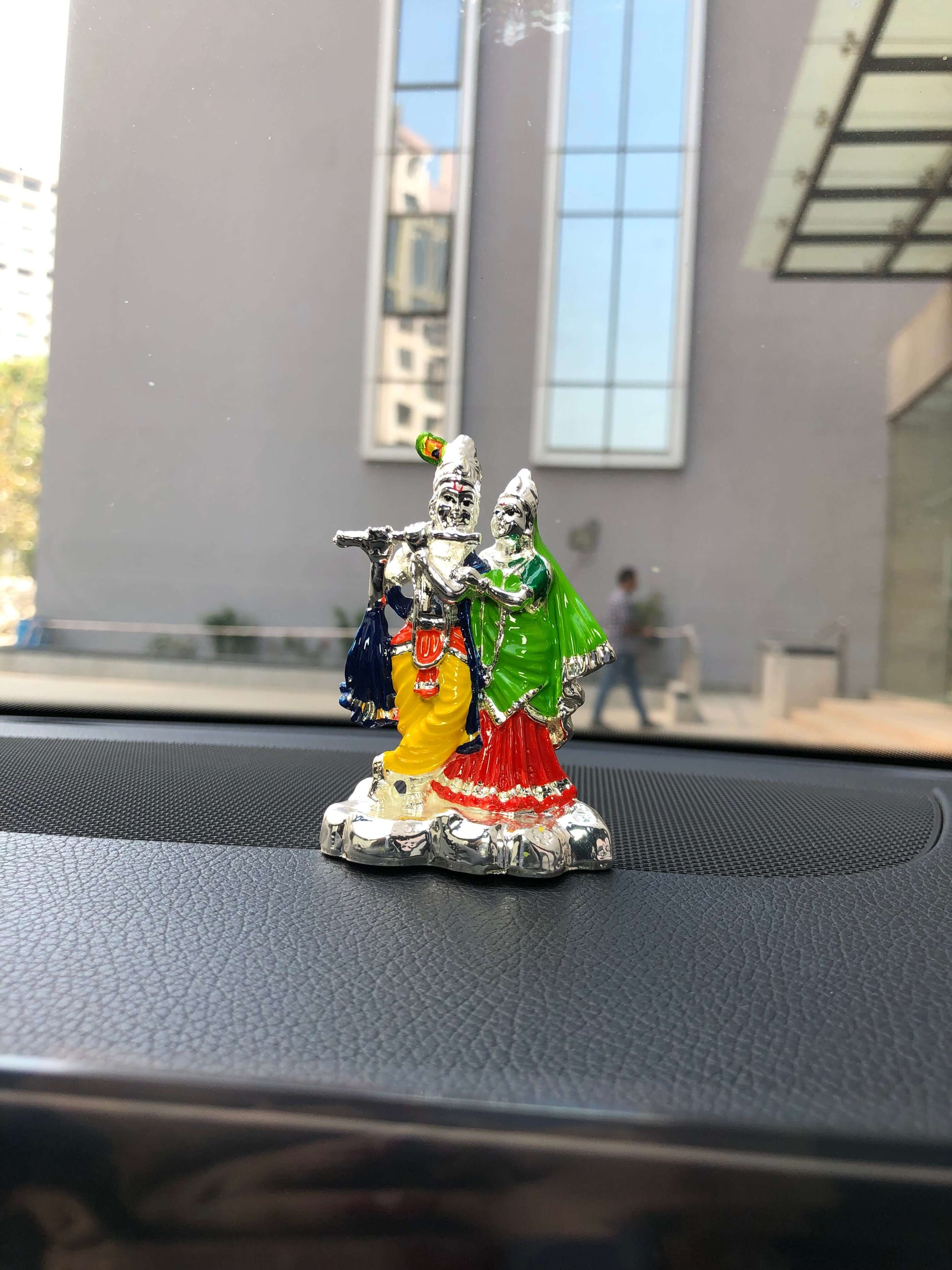 A colorful Radha Krishna idol, possibly silver-plated and painted, sits on the dashboard of a car with a modern building visible through the windshield in the background. The idol depicts Krishna playing the flute and Radha standing beside him.
