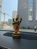 A close-up shot of a dark-colored, gold-plated Lord Balaji (Venkateswara) idol placed on the dashboard of a car, with a blurred cityscape visible through the windshield in the background.