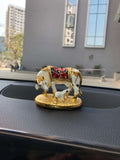 Golden Kamadhenu cow and calf idol with colorful accents displayed on a car dashboard.
