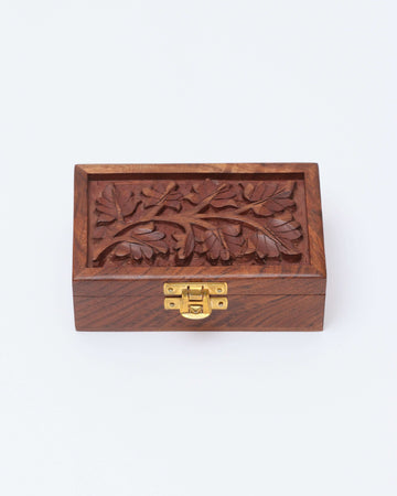 A small, brown wooden box is shown with a brass latch on the front. The lid of the box features a handcrafted floral engraving of leaves and branches.
