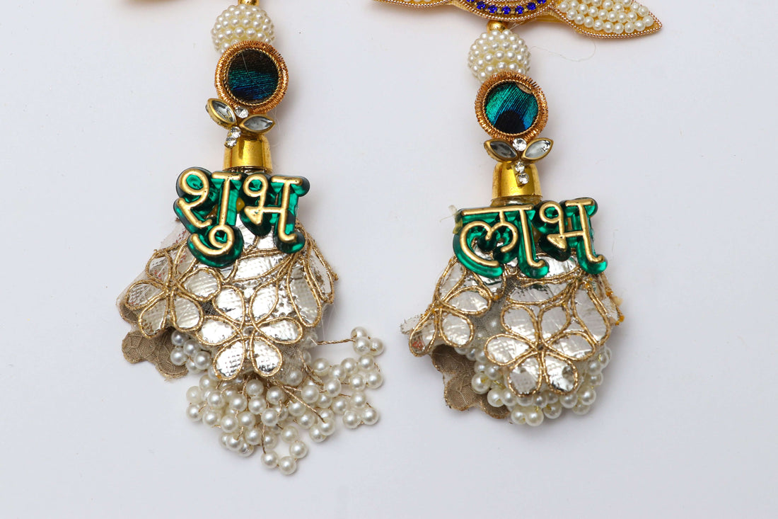 A pair of decorative Shubh Labh door hangings, featuring a beaded lotus design with a small peacock motif and a circular yellow backing. Below the lotus, a dangling silver bell-shaped piece is decorated with a green text "शुभ" and "लाभ" respectively, and small pearls hanging from the bottom.