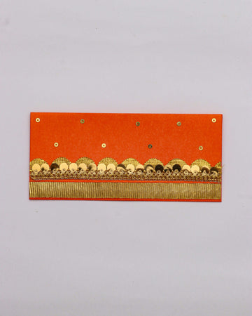An orange Shagun envelope with gold and black embellished trim and a fringe of gold thread along the bottom. The envelope is also decorated with small gold sequins scattered across the top half.