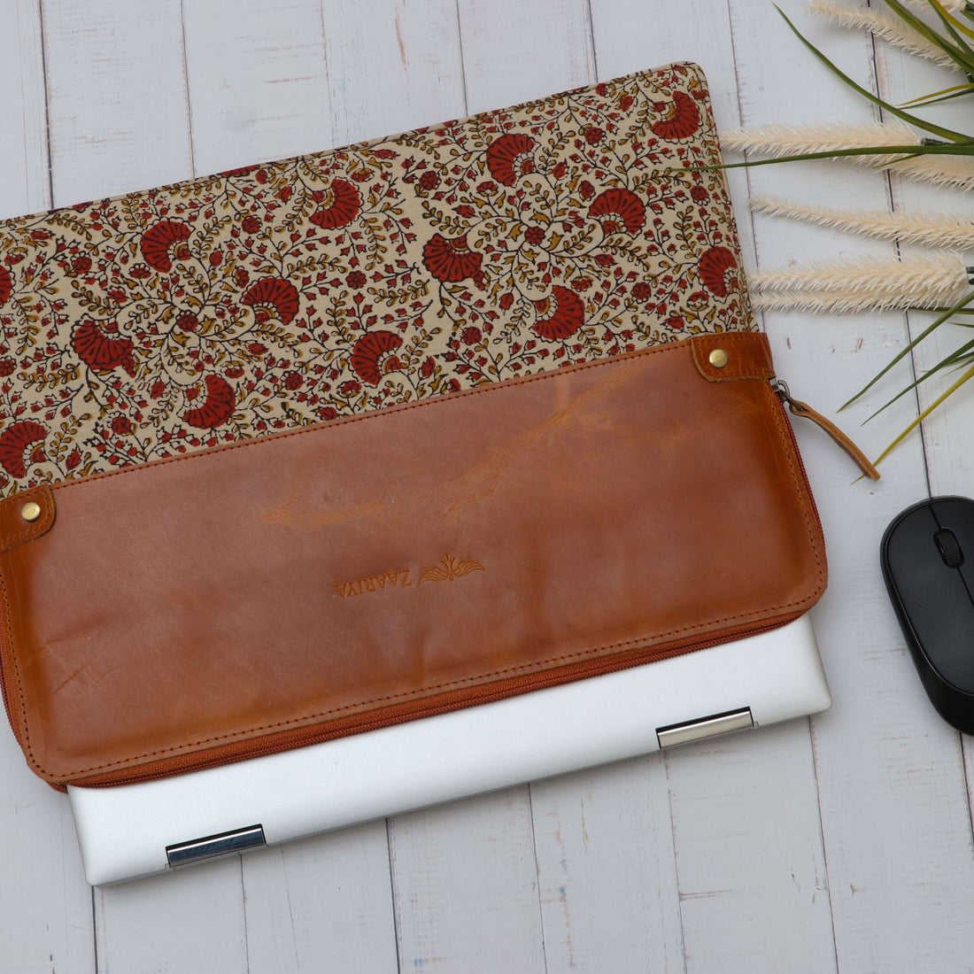 A brown leather laptop sleeve with floral block print design, displayed alongside a white laptop and a computer mouse on a wooden surface.