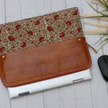 A brown leather laptop sleeve with floral block print design, displayed alongside a white laptop and a computer mouse on a wooden surface.