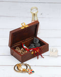 A small, dark wooden jewelry box with a brass latch is open, revealing various pieces of jewelry inside, including gold-toned chains, beaded necklaces, and black earrings. In the foreground, a pair of gold hoop earrings and a rectangular pendant with a colorful geometric pattern are visible. A perfume bottle stands in the background on a distressed white wooden surface.