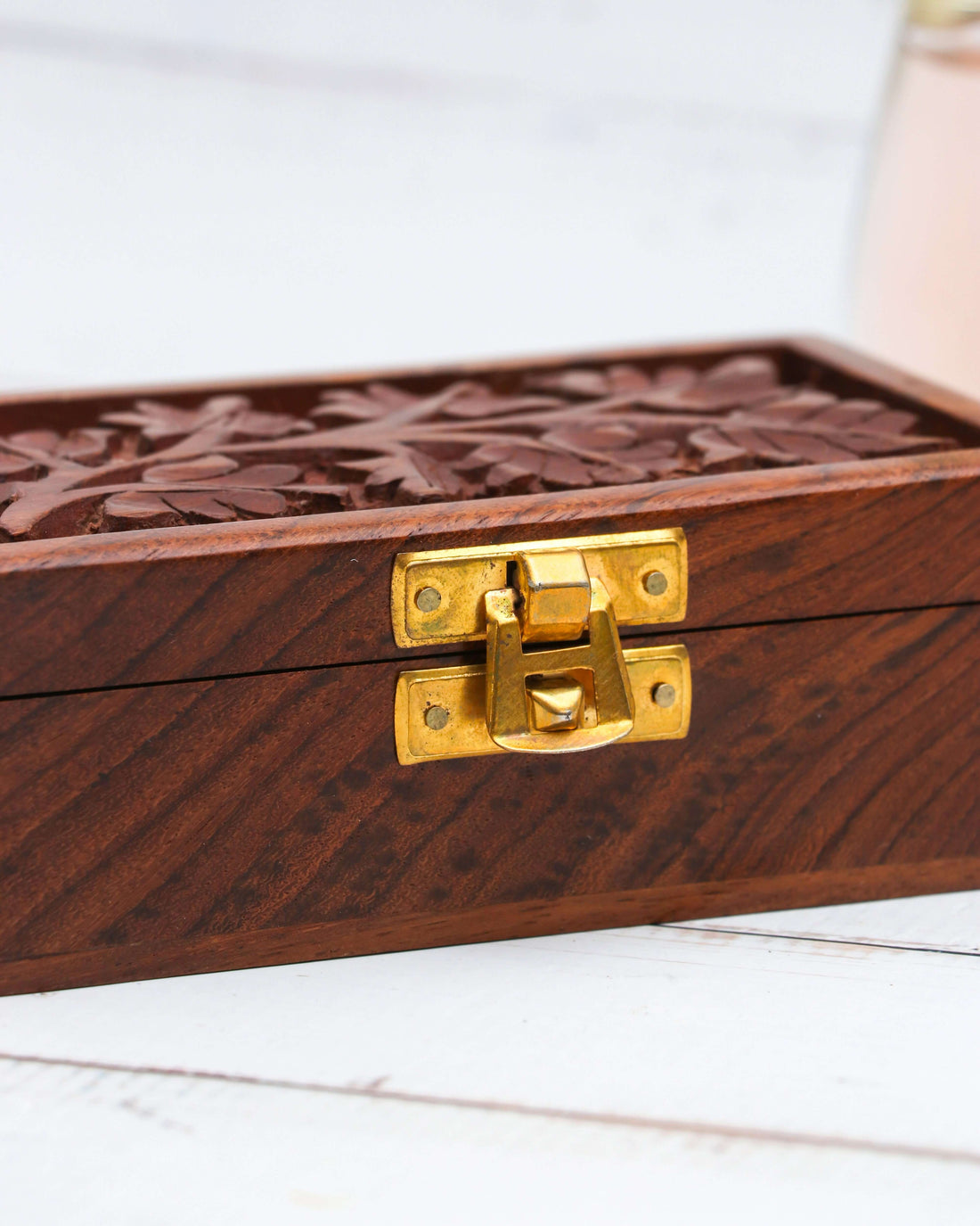 A small, brown wooden box is shown with a brass latch on the front. The lid of the box features a handcrafted floral engraving of leaves and branches.