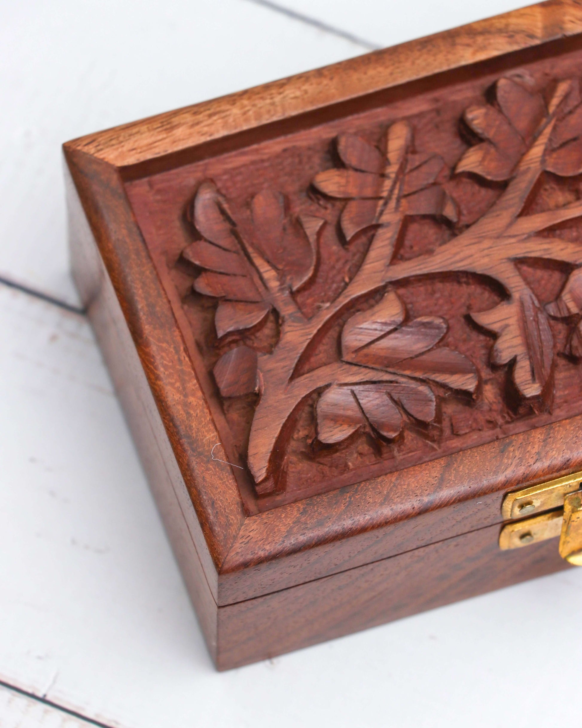 A small, brown wooden box with a gold-colored latch on the front. The lid of the Handcrafted Floral Engraving Jwellery Box features an intricate carving of a leafy tree design.