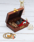 An open, handcrafted wooden jewelry box with brass hardware, showcasing various pieces of jewelry inside. The box contains a gold-toned necklace, black earrings, and red and white beaded earrings. Additional jewelry, including a gold chain and bangles, is visible on the white wooden surface in front of the box.