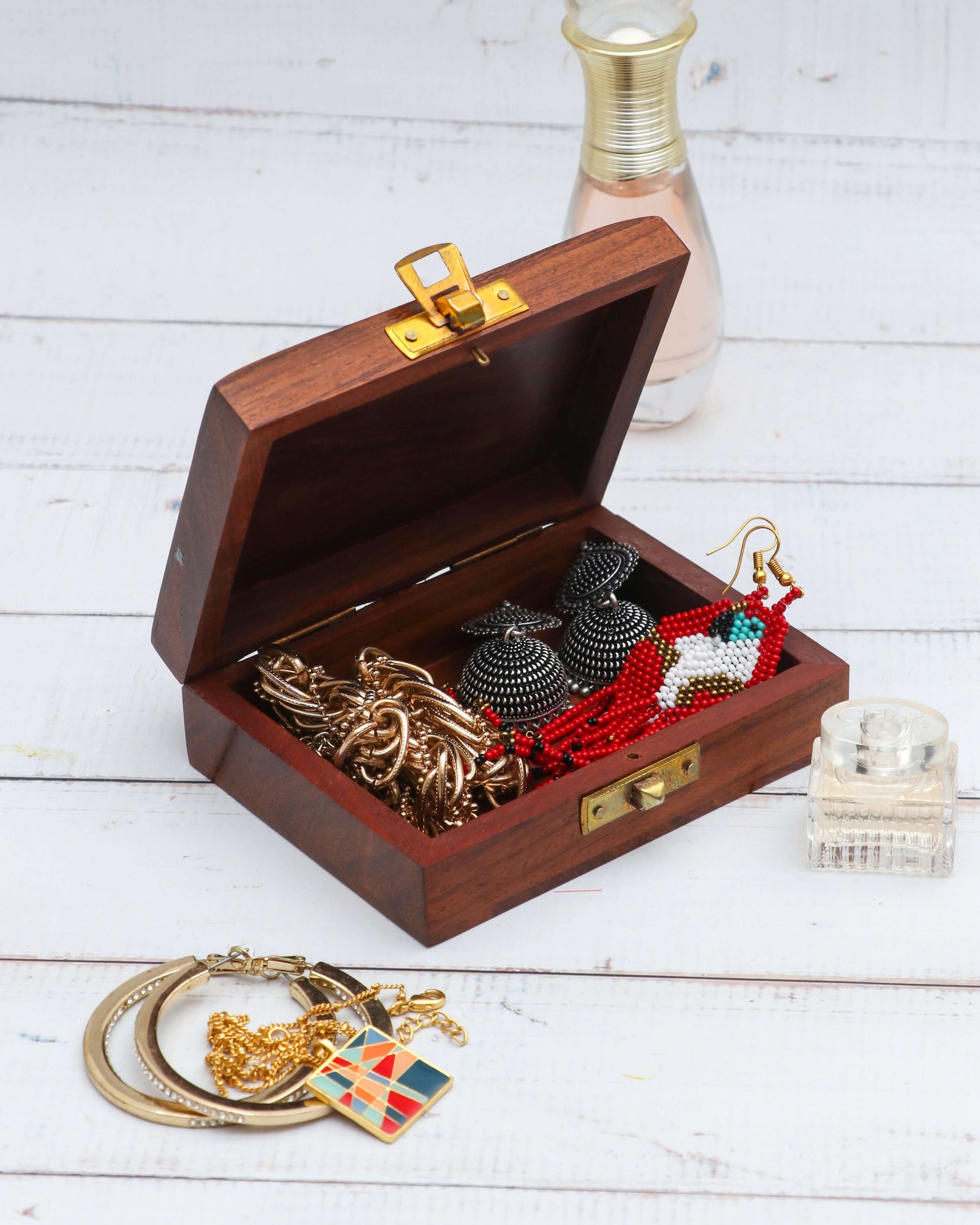 An open, handcrafted wooden jewelry box with brass hardware, showcasing various pieces of jewelry inside. The box contains a gold-toned necklace, black earrings, and red and white beaded earrings. Additional jewelry, including a gold chain and bangles, is visible on the white wooden surface in front of the box.