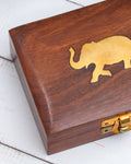 A small, rectangular, handcrafted wooden jewelry box with a brass latch on the front and a brass elephant inlaid on the lid. The wood has a natural grain and a polished finish.