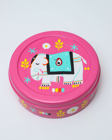 A pink, round hand-painted steel spice box featuring a white elephant with a turquoise-colored blanket on its back. The design also includes yellow leaves, white flowers, and small dot patterns.