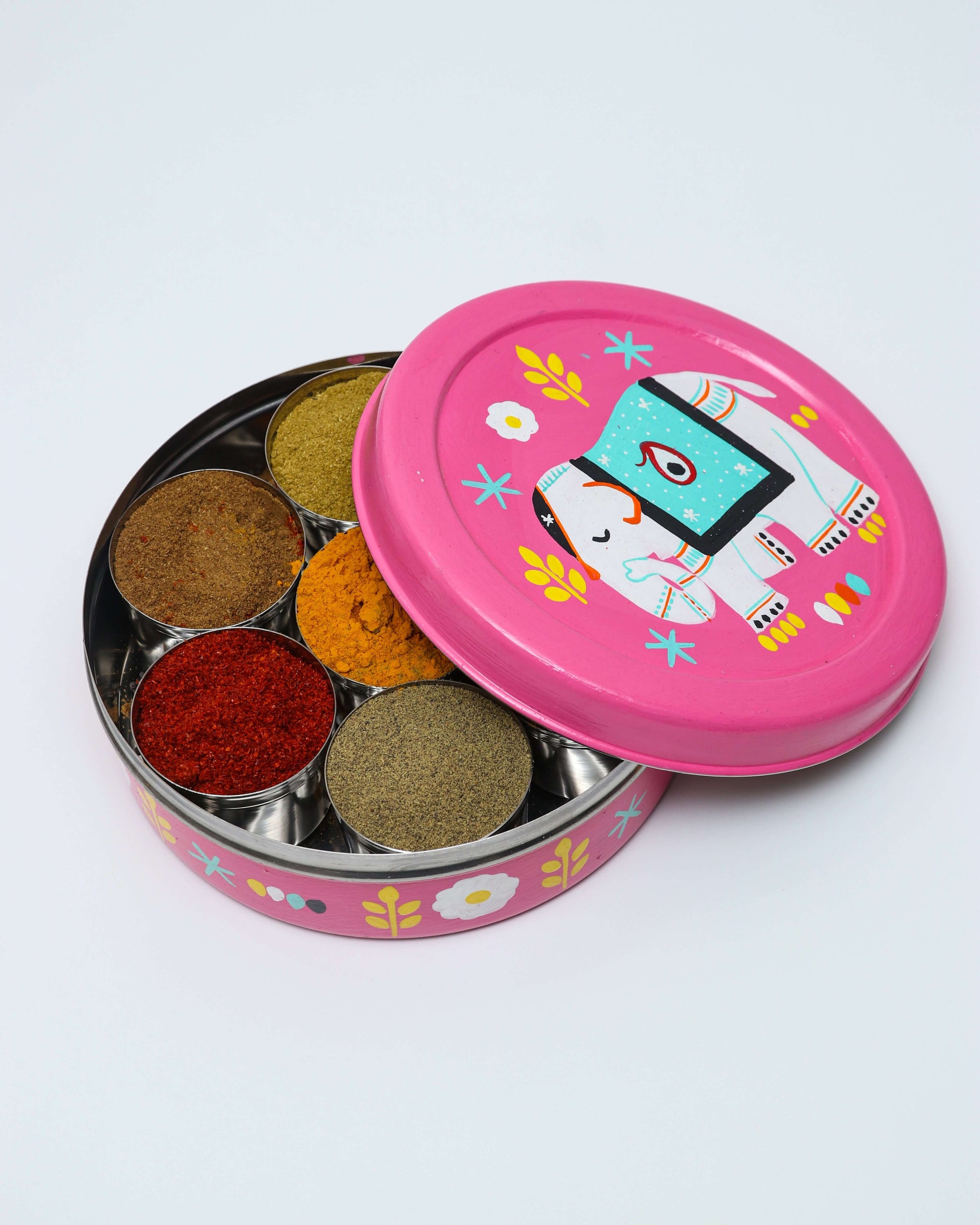A round, pink hand-painted steel spice box with six individual compartments filled with different colored spices. The lid features a white elephant with a decorative turquoise and blue blanket on its back, surrounded by yellow leaves and white flowers. The side of the spice box is also decorated with similar floral patterns. The lid is shown partially open, resting next to the container.