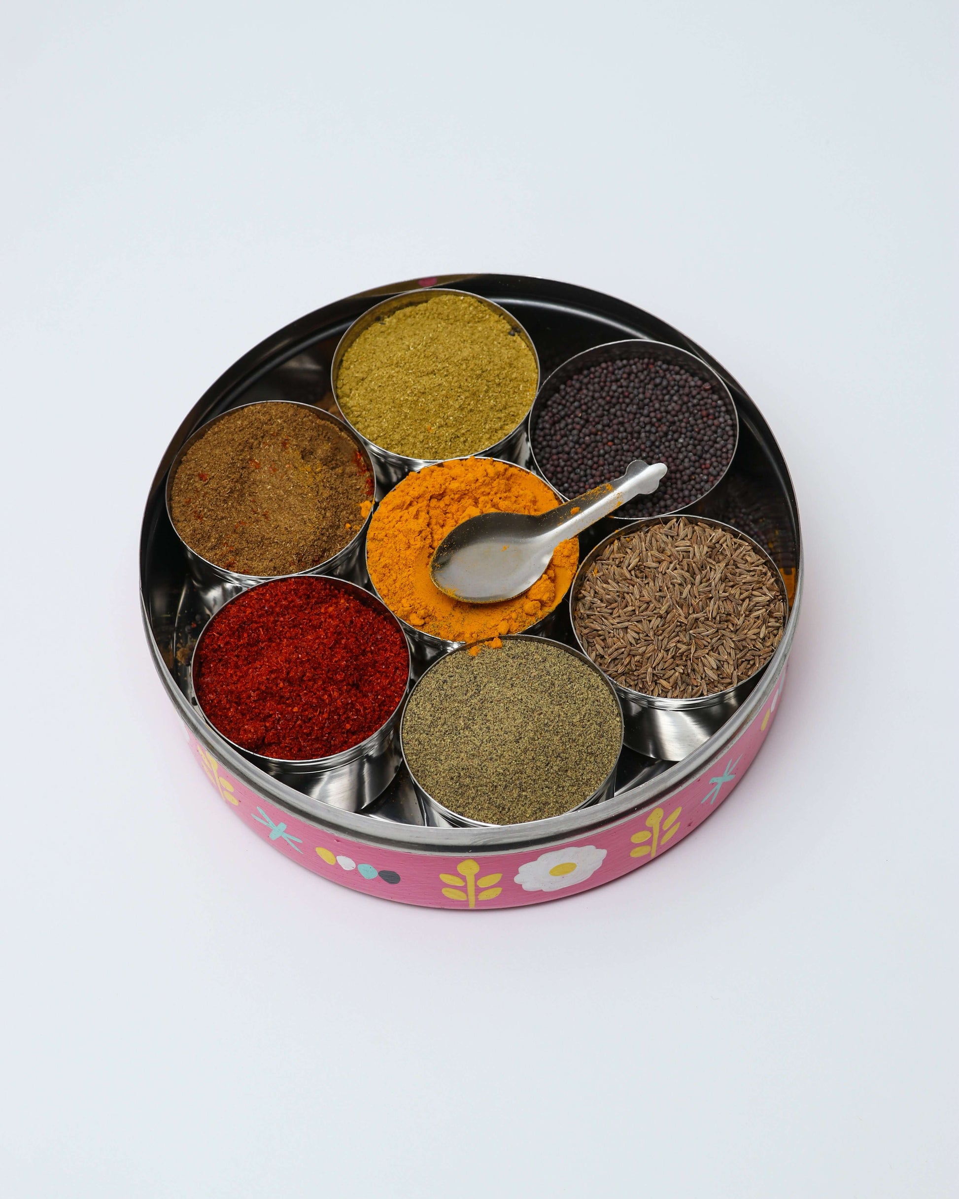 A round, steel spice box, or masala dabba, with a pink, hand-painted floral pattern on the exterior. The box is open, revealing seven compartments filled with a variety of colorful spices, including a bright orange-yellow powder and dark brown seeds. A small metal spoon rests inside one of the compartments.