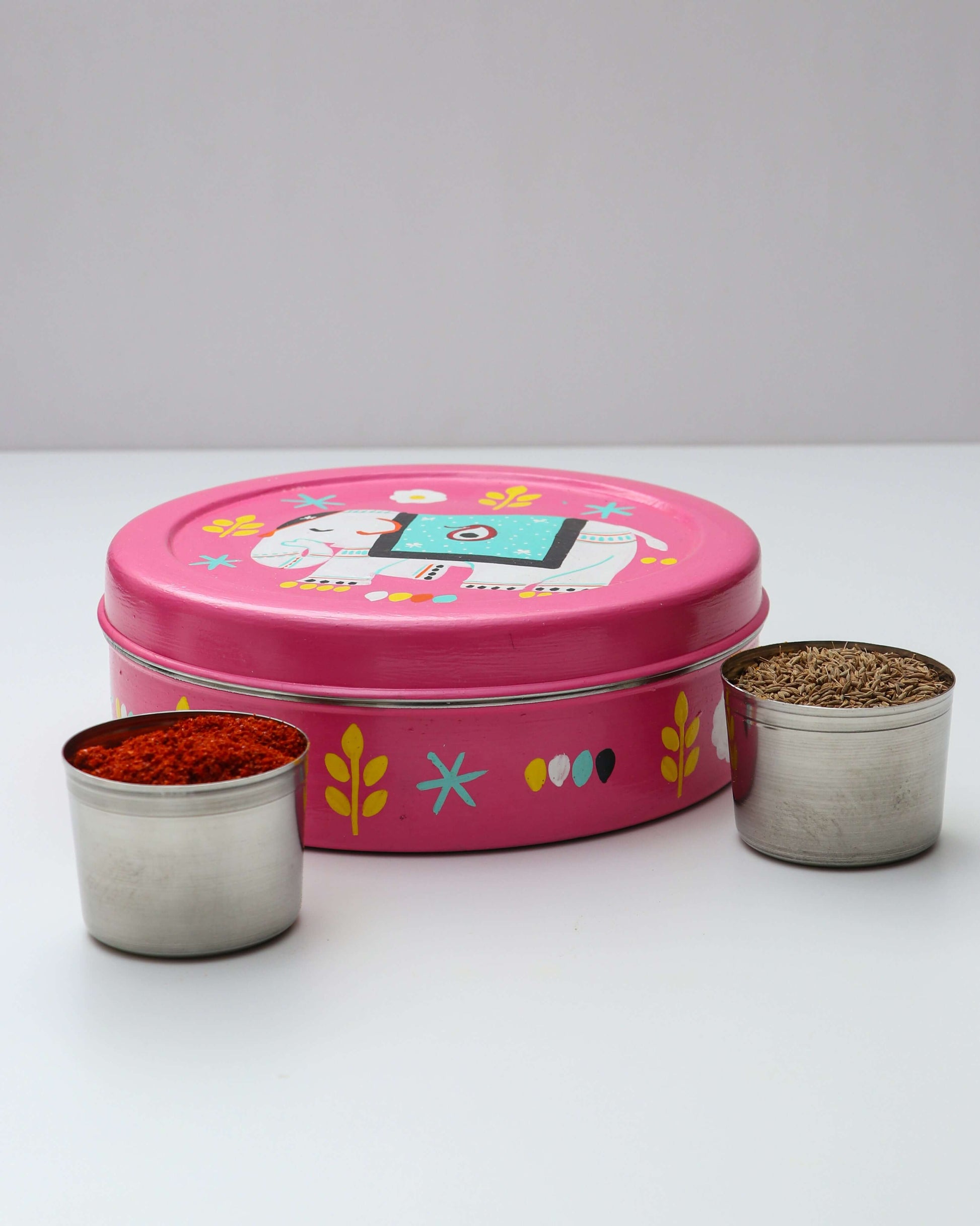 A round, pink hand-painted steel spice box with a lid featuring a white elephant with a blue blanket, yellow leaves, and white flowers. Two small steel containers, one filled with red powder (chili powder) and the other with brown seeds (cumin), are placed on either side of the box.