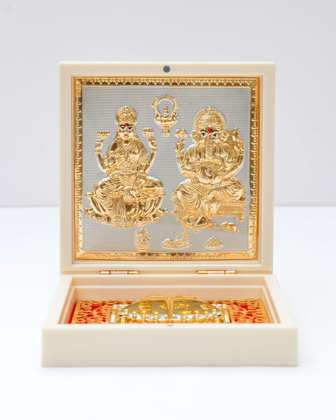 A white decorative box containing golden idols of Lakshmi and Ganesha, placed amidst marigold flowers, rose petals, traditional Indian sweets (ladoos), and brass diyas, likely set up for a Hindu festival or puja celebration.