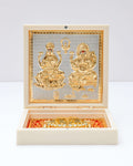 A decorative white and gold box open to reveal golden embossed idols of Lakshmi and Ganesha, with gold charan padukas (footprints) at the bottom.