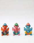 A row of three small, handpainted statues of the Hindu deity Lord Ganesha is displayed against a white background. The statues, which appear to be made of wood or ceramic, are seated in a similar pose. From left to right, the first Ganesha is orange with a red-and-blue hat and holds a musical instrument; the second is pink with a green-and-white hat; and the third is blue with a red-and-gold hat and is holding cymbals. Each statue is decorated with a different colorful pattern.