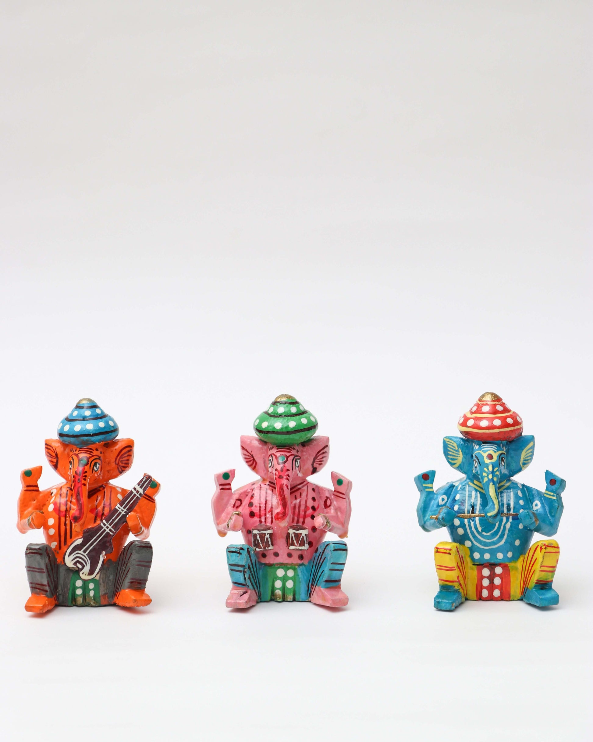 A row of three small, handpainted statues of the Hindu deity Lord Ganesha is displayed against a white background. The statues, which appear to be made of wood or ceramic, are seated in a similar pose. From left to right, the first Ganesha is orange with a red-and-blue hat and holds a musical instrument; the second is pink with a green-and-white hat; and the third is blue with a red-and-gold hat and is holding cymbals. Each statue is decorated with a different colorful pattern.