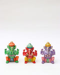 A set of three hand-painted, seated Wooden Ganesh Statue Products is displayed in a row on a white background. The statues are vibrantly colored: one is green and orange, the second is red and yellow, and the third is purple and pink. Each Ganesha figure wears a unique, striped hat and a distinct, detailed costume with small white and yellow dot patterns.
