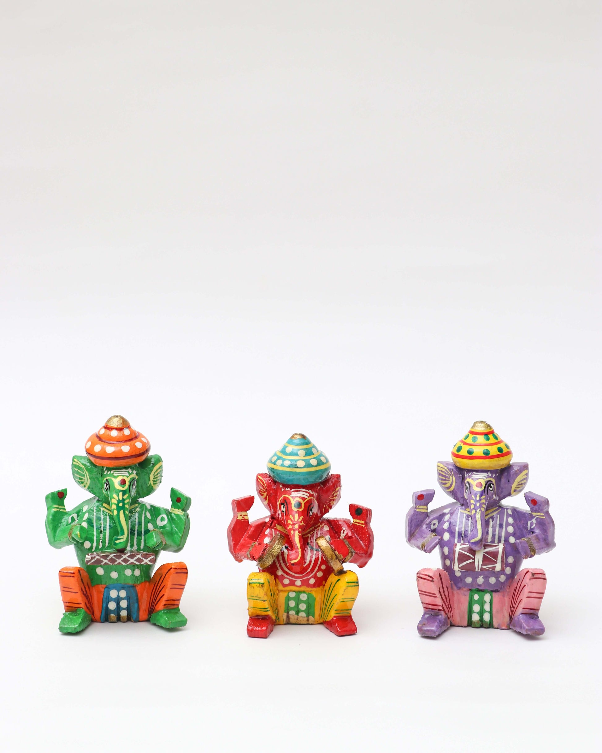 A set of three hand-painted, seated Wooden Ganesh Statue Products is displayed in a row on a white background. The statues are vibrantly colored: one is green and orange, the second is red and yellow, and the third is purple and pink. Each Ganesha figure wears a unique, striped hat and a distinct, detailed costume with small white and yellow dot patterns.