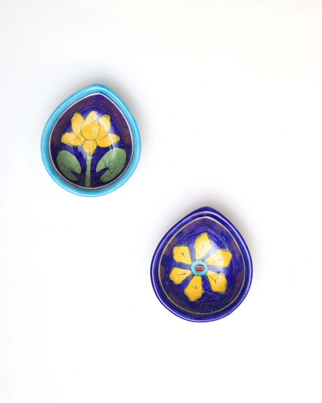 A pair of tear-drop-shaped, dark blue Assorted Handpainted Ceramic Diya is displayed on a white background. One diya features a vibrant yellow lotus with green leaves, while the other is decorated with a yellow star-like flower and a red center.