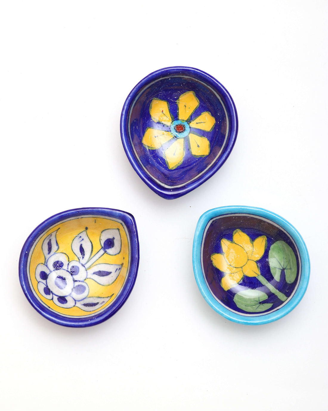 A pair of tear-drop-shaped, dark blue Assorted Handpainted Ceramic Diya is displayed on a white background. One diya features a vibrant yellow lotus with green leaves, while the other is decorated with a yellow star-like flower and a red center.