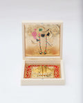 Golden framed picture of Shreenathji and Charan Paduka in a white wooden box with red fabric lining.